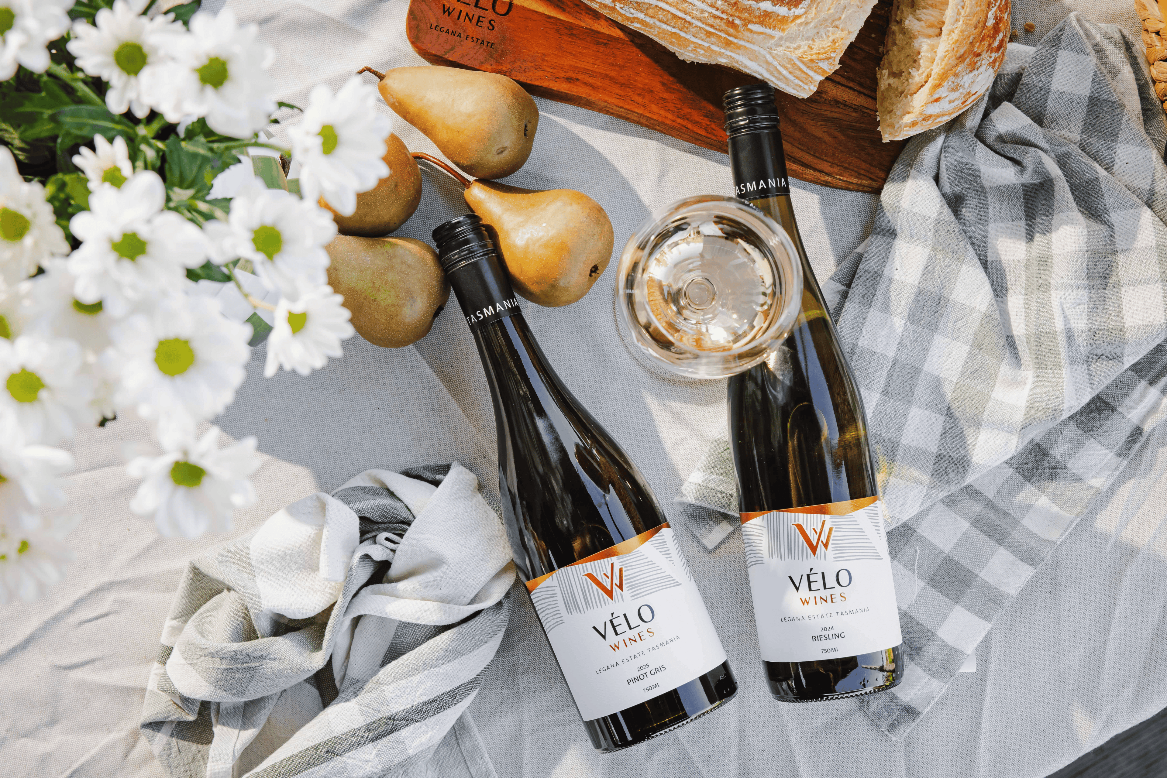 Two bottles of Velo Winery wine arranged on a white picnic blanket with a glass of white wine, pears, white daisies, bread, and a plaid cloth.