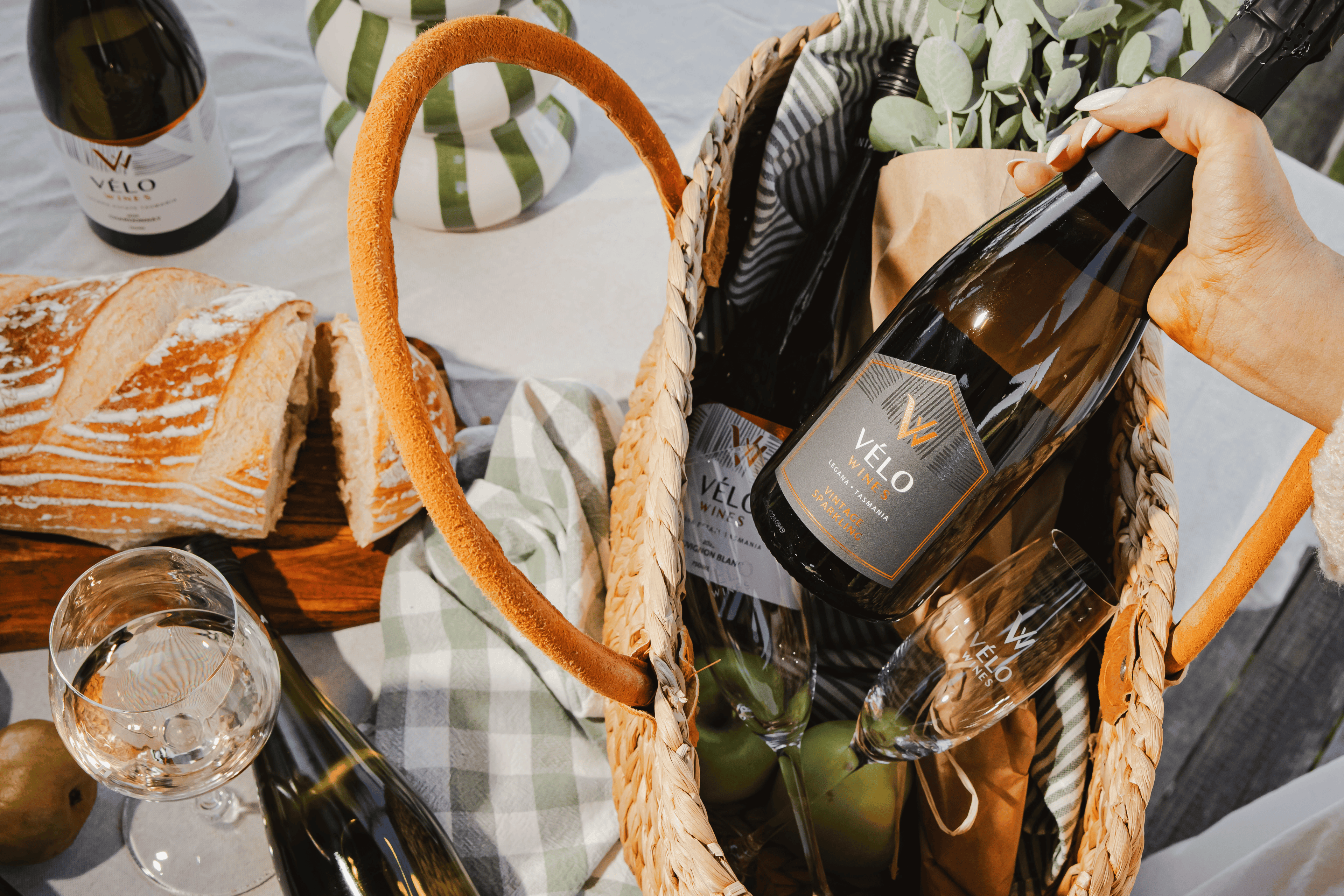 A hand holds a bottle of Velo white wine inside a woven straw picnic basket, surrounded by bread, wine glasses, and a checkered cloth on a white tablecloth.